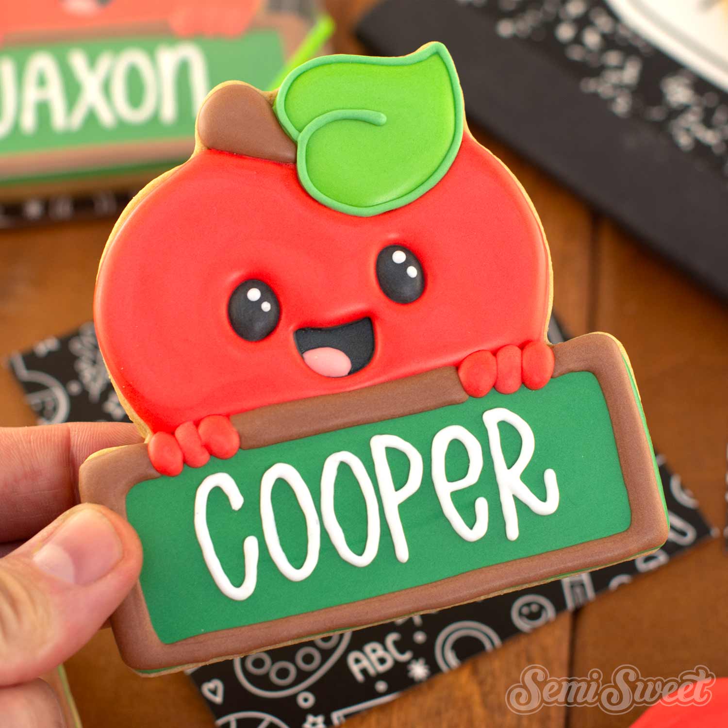 A hand holds a decorated sugar cookie shaped like a smiling red apple with a green leaf and a green sign that says "COOPER" in white letters. The background shows a wooden table and other themed cookies or packaging.