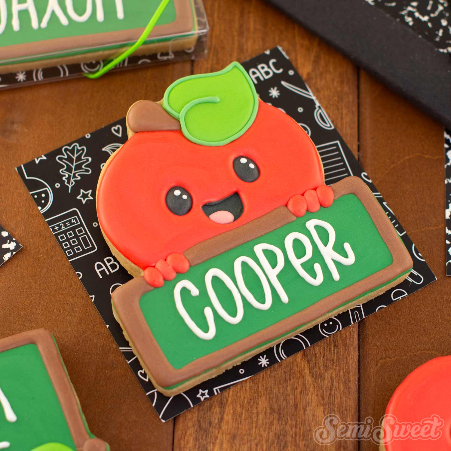 A decorated sugar cookie shaped like a smiling red apple with a green leaf, holding a green sign that says 'COOPER' in white letters. The cookie is placed on a black patterned card on a wooden table, with other similar cookies partially visible around it.