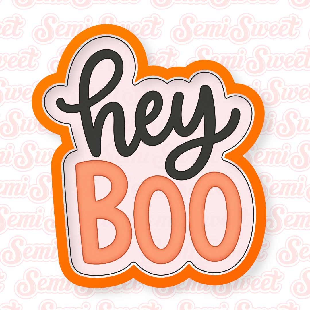 A cookie cutter with the words 'hey BOO' in stylized lettering. The top word 'hey' is in dark charcoal script, and the bottom word 'BOO' is in peach block letters, all on a pale pink background with a thick orange border.